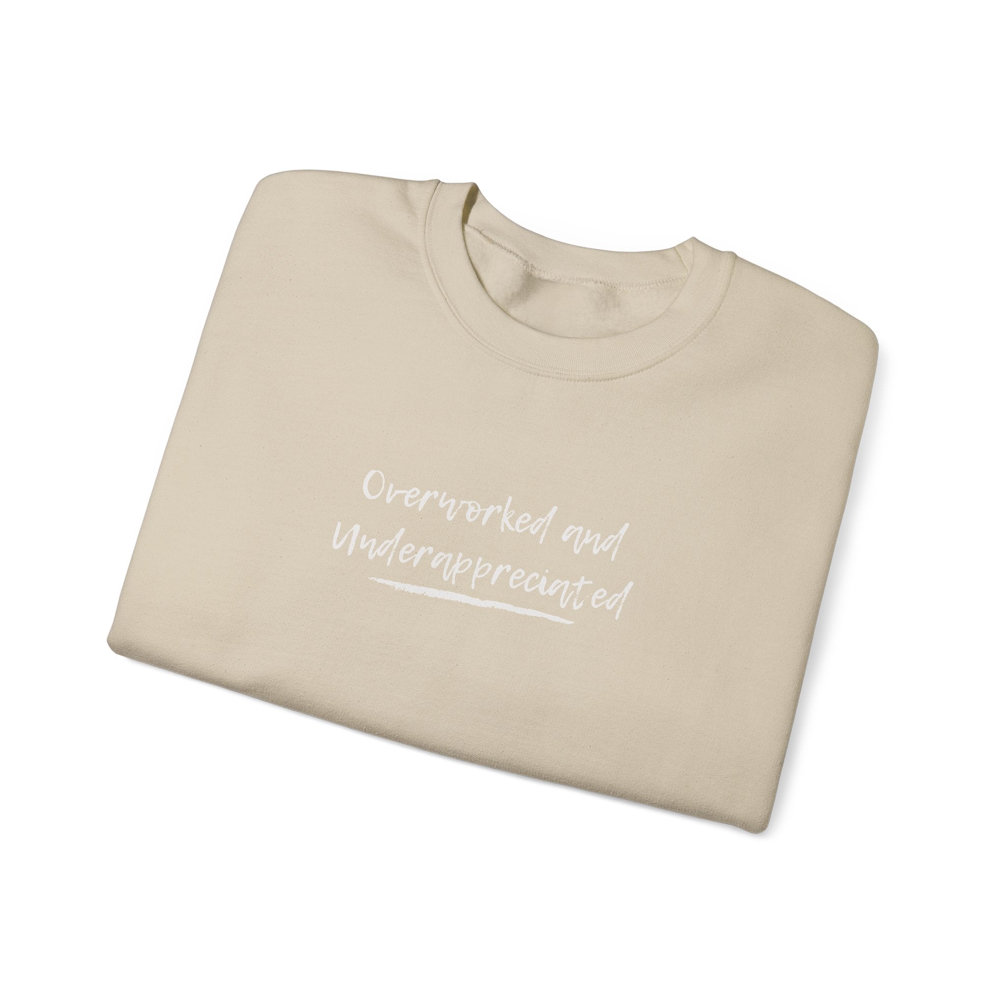 Beige folded sweatshirt with 'overworked and underappreciated' text and heart symbol on a white background