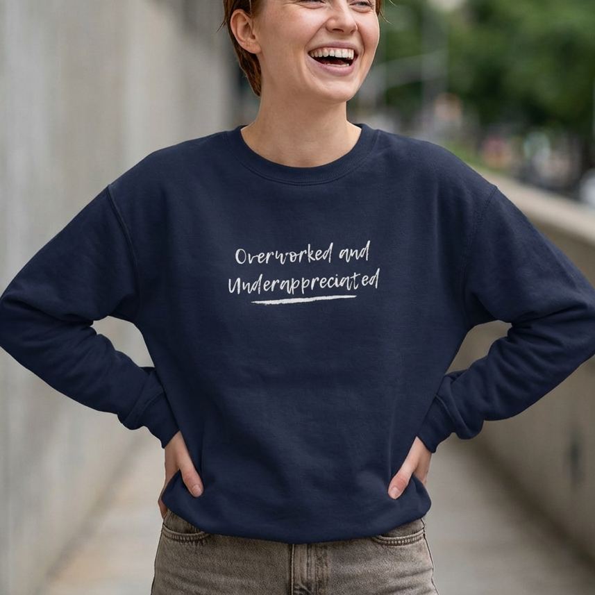 Person wearing a navy blue sweatshirt with 'Overworked and Underappreciated' text, standing outdoors. perfect for burnout gift