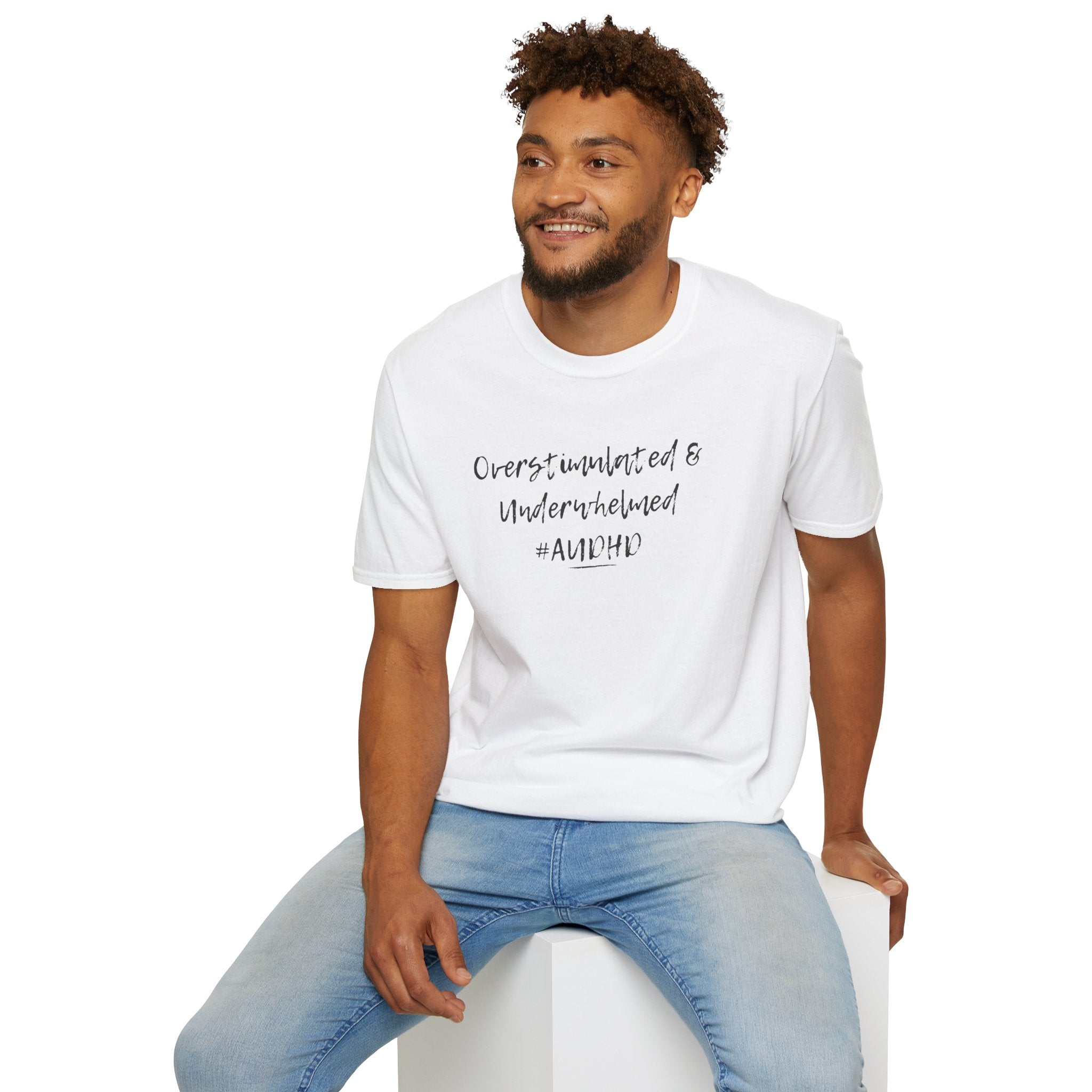 Man wearing a white t-shirt with  "Overstimulated" text. Designed for autistic and ADHD individuals with sensory processing sensitivities to prevent tactile irritation. sitting on a white stool against a white background.